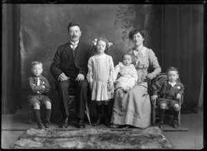 Studio family unidentified portrait, husband with moustache and wife sitting with baby daughter and young twin sons, older daughter in pigtails and bows standing, Christchurch
