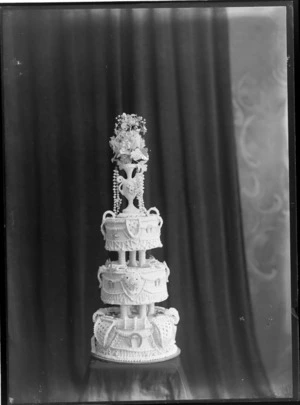 Three tiered wedding cake with elaborate icing on sides, columns and vase with flowers on top, probably Christchurch region