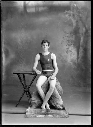 Studio portrait of unidentified young sportsman in costume with medal, sitting on fur rug with table, Christchurch