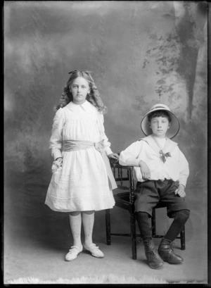 Studio portrait of unidentified young girl and boy, shows boy wearing a hat and sitting on a chair, probably Christchurch district