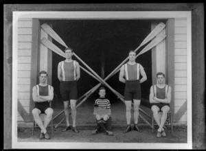Group portrait of members of the Avon Rowing Club, showing four young men in swimming costumes, with a young boy coxswain sitting in center [holding a boat rudder?], and oars crossed diagonally behind, outside the Avon Rowing Club boatshed, Christchurch