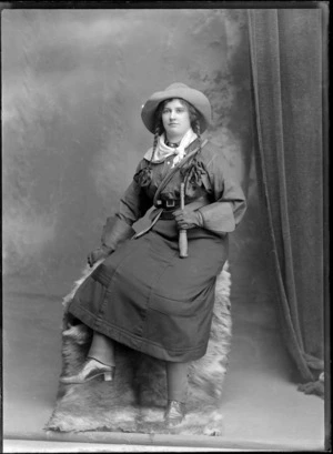 Studio portrait of an unidentified young woman, wearing a 'Wild West' costume, including leather gauntlets, gun holster, a handkerchief tied around neck, plaited pigtails, and a whip across chest, possibly Christchurch district