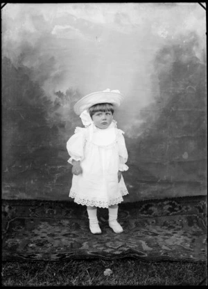 Studio portrait of an unidentified small girl, wearing a wide-brimmed hat tied under chin, a lace-trimmed bib, possibly Christchurch district