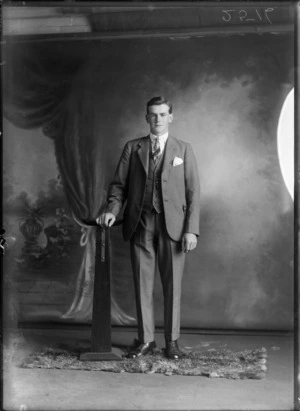 Studio portrait of unidentified young man in three piece suit, striped shirt and tie, with greenstone watch chain pendant, standing with wooden font, Christchurch