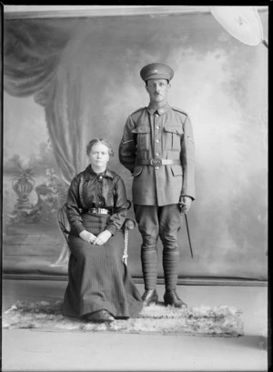 Studio unidentified family portrait, a Lance Corporal World War I soldier with toothbrush moustache, [Tui or Huia?] collar and hat badges and swagger stick, standing alongside wife in black lace blouse with brooch, Christchurch