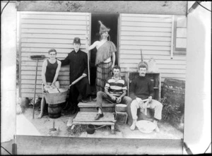 Unidentified group of men performing domestic chores outdoors, shows some men peeling potatoes, cleaning and washing dishes, also includes a man dressed in Maori costume holding a stick, probably Christchurch district