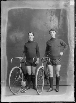 Studio portrait of two unidentified males, with racing bicycles, trophy and badges, probably Christchurch district