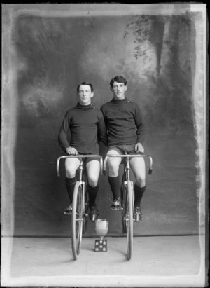 Studio portrait of two unidentified males, sitting on racing bicycles, with cup trophy and badges, probably Christchurch district