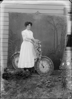 Unidentified woman outside a wooden house, with a bicycle decorated with flowers, and a painted studio backdrop, probably Christchurch district