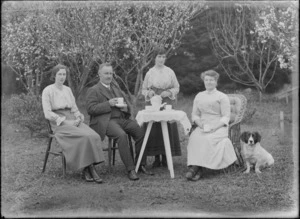 Unidentified man and three women having tea outdoors, shows table set with cups and saucers, and a young woman holding a teapot, also includes a dog, probably Christchurch district