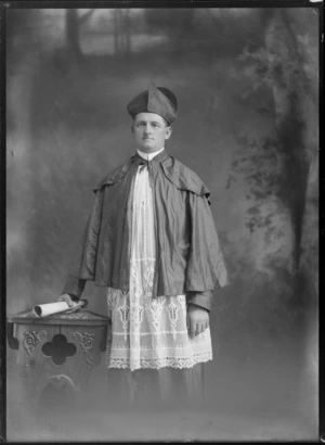 Studio portrait of unidentified man, [a Bishop?] in religious attire, standing by a carved wooden table, probably Christchurch district
