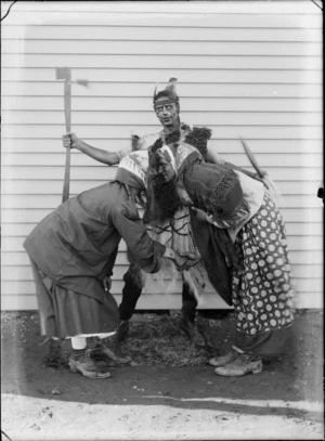 Group of three unidentified men in 'native' costume, outside a building, man in the background holding a stick, [two men in foreground performing a hongi?], probably Christchurch district