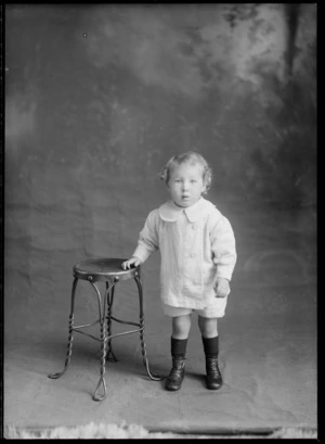 Studio portrait of unidentified small child, standing next to a stool, probably Christchurch district