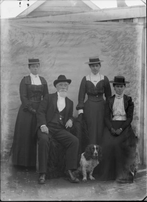 Group of three unidentified women and an elderly man, with a dog, outdoors, with a painted studio backdrop, probably Christchurch district