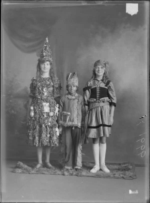 Studio portrait of two unidentified girls and a boy, all dressed in fancy dress costumes, shows the boy dressed in a 'Red Indian' costume, and one girl dressed as a Christmas tree, probably Christchurch district