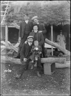 Group of four unidentified adult males some wearing 'silly' hats, and a small boy, outside a hut with a thatched roof, probably Christchurch district, includes two unidentified women in the background