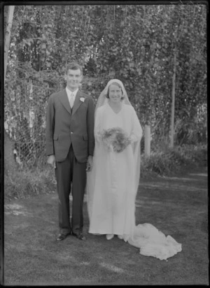 Outdoors on grass in front of trees, an unidentified wedding couple portrait, bride with extra long veil and pearl necklace holding flowers standing with the groom, probably Christchurch region