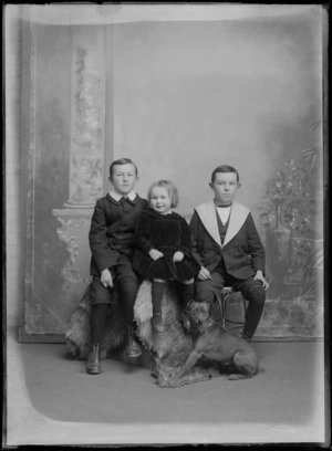 Studio unidentified family portrait of three young children sitting, older brother in a large collar double breasted jacket, younger in a coat and Eton shirt collar, with a young sister between them, dog in front, Christchurch