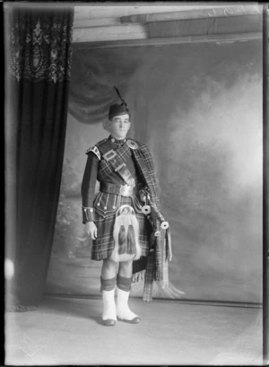 Studio portrait of an unidentified young Scottish piper in full costume, with collar and shoulder badges, Glengarry hat with feather, sporran, sash and bagpipes, Christchurch