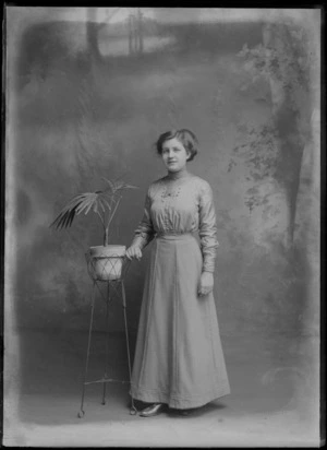 Studio portrait of an unidentified young woman in a pleated high neck collar blouse with brooch and matching skirt, standing with a potted plant with stand, Christchurch