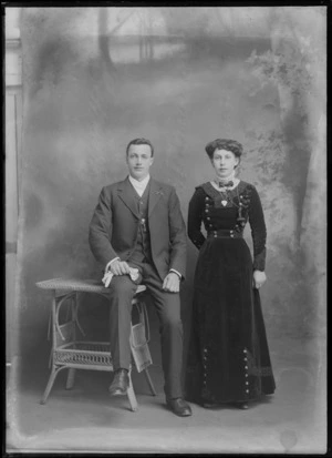 Studio portrait of an unidentified couple, man with waistband pendant and white bow tie, woman in a double breasted dark velvet dress with sleeve and neck pattern, a lace collar with bow, bar brooch and heart shaped necklace, Christchurch