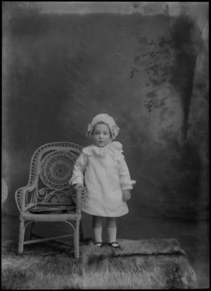 Studio unidentified family portrait of a young girl in a lace collar and sleeve corduroy dress and bonnet with bow, standing next to a cane chair, Christchurch
