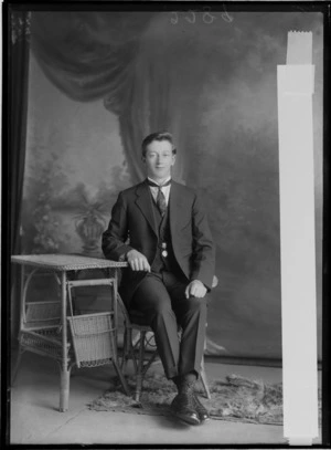 Studio portrait of an unidentified young man in a three piece suit, dark patterned tie and waistcoat band pendant sitting with a cane table, Christchurch