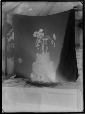 Outdoors within a wedding reception tent in front of a hand held black cloth, three tiered wedding cake with lucky horse shoes topped with a vase and flowers, probably Christchurch region