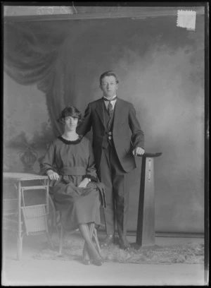 Studio portrait of an unidentified young couple, man in a three piece suit, patterned tie and waistcoat band pendant standing with a wooden font, woman in a fur collar and sleeve lined dark dress with wrist watch sitting with a cane table, Christchurch