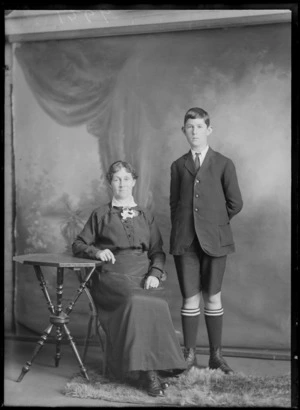 Studio unidentified family portrait, mother sitting with flower carnation and older son in pin striped jacket and shorts, shirt and tie with school socks standing, Christchurch