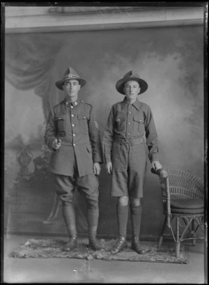 Studio family portrait, unidentified World War I Sergeant with collar and hat badges holding a swagger stick standing alongside a young man in soldier's jacket, shorts and hat, Christchurch