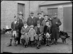 Outdoors behind brick building, unidentified group portrait of eight men and two boys, with a man and boy in Scottish costumes with piper's sporrans, probably Christchurch region