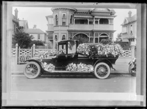 Outdoors view of a truck carrying an unidentified coffin and covered in flowers, on the street in front of a two story urban wooden building with verandas, probably Christchurch region