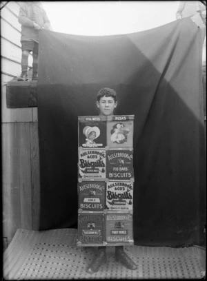 Outdoors portrait in front of a dark sheet, an unidentified teenaged boy, head uncovered, in fancy dress 'Aulsebrook & Co' biscuit boxes costume, probably Christchurch region