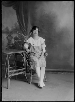 Studio portrait of an unidentified young woman in a large white collar striped dress with a flax braided belt, bar and figurine brooch, sitting with a cane table, Christchurch