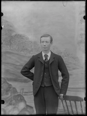 Outdoor in front of a false backdrop, a portrait of an unidentified older teenaged boy in a three piece suit, striped shirt and large dark tie, standing with a wooden chair, probably Christchurch region