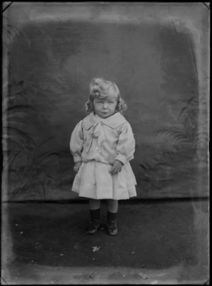 Outdoor in front of a false backdrop, an unidentified family portrait of a young girl in a large collar shirt with a bow and matching pleated skirt standing, probably Christchurch region