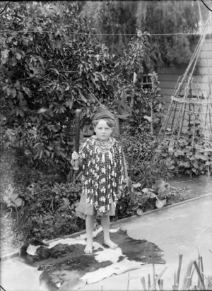 Outside unidentified portrait in the back garden, a young boy dressed up in a beaded cloak over a striped shirt and hat, holding a small bat, probably Christchurch region