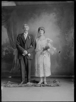 Studio unidentified wedding couple portrait, groom with large moustache, butterfly shirt collar and three piece suit with lapel flowers, bride with long veil and embroidered dress holding flowers and lucky horse shoe, Christchurch