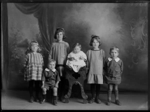 Studio unidentified family portrait of five young children grouped around a baby with a large crochet bib on a wooden highchair, with two girls and two boys in identical clothes, Christchurch
