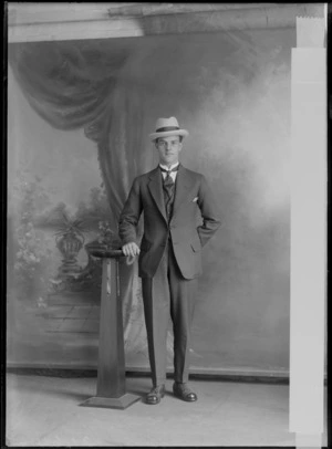 Studio portrait of an unidentified young man in a pinstriped suit, butterfly shirt collar and patterned tie with tie pin and hat standing next to a wooden font, Christchurch
