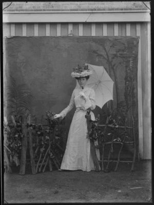 Outdoors in front of a false backdrop, portrait of an unidentified young woman in a high lace neck blouse and skirt with a flower pattern, brooches and corsage, standing in a flower hat holding an open umbrella beside a fence prop, probably Christchurch region