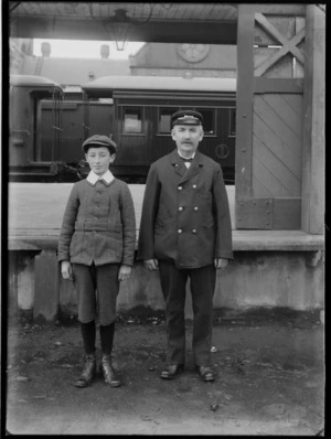 Outdoor in a loading bay of a railway station with a train behind, portrait of an unidentified older man with a moustache in a railway worker's uniform standing with an older boy with an Eton shirt collar, a cloth cap, tweed coat and pants, probably Christchurch region