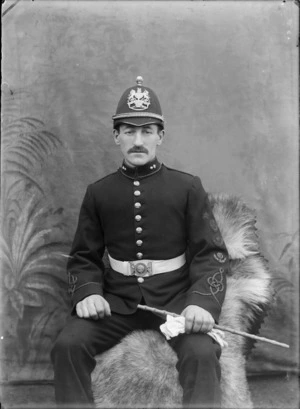 Outdoors in front of false backdrop, portrait of an unidentified soldier in dress uniform, with moustache, collar badges, sleeve insignia, wearing a domed helmet with large lion and unicorn badge, probably Christchurch region