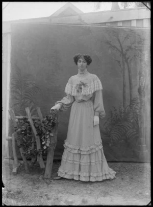 Outdoors in front of trees, portrait of an unidentified young woman in a high neck lace patterned dress, corsage, gloves, bar brooch, pearl necklace and heart shaped pendant standing with a fence prop, probably Christchurch region