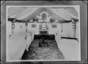 Inside a large tent with Trivoli Camp sign set up as a bedroom with carpet, six single beds, a dresser with mirror, a small table, posters and framed pictures, [Sumner?], Christchurch