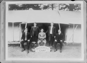 Outdoors in front of two overlapping large tents, five unidentified young men in suits and double round, arundel and butterfly shirt collars, with potted plants and 'Bluebell Camp' sign, [Sumner?], Christchurch