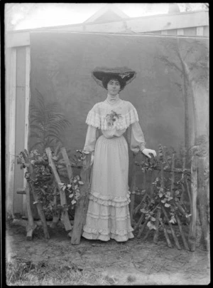 Outdoors in front of trees, portrait of an unidentified young woman in a high neck lace patterned dress, corsage, gloves, bar brooch, pearl necklace and heart shaped pendant, standing in a large lace dark hat with a fence prop, probably Christchurch region