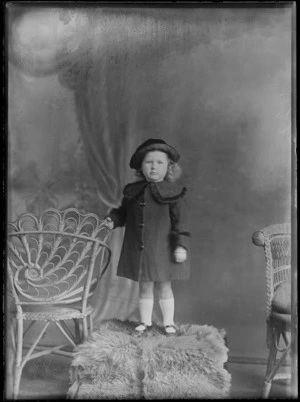 Studio unidentified family portrait of a young girl in a large embroidered fur lined collar dark coat with large buttons, hat and white socks with leather shoes, standing on a fur rug covered box next to cane chair, Christchurch