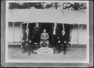 Outdoors in front of two overlapping large tents, five unidentified young men in suits and double round, arundel and butterfly shirt collars, with potted plants and 'Bluebell Camp' sign, [Sumner?], Christchurch